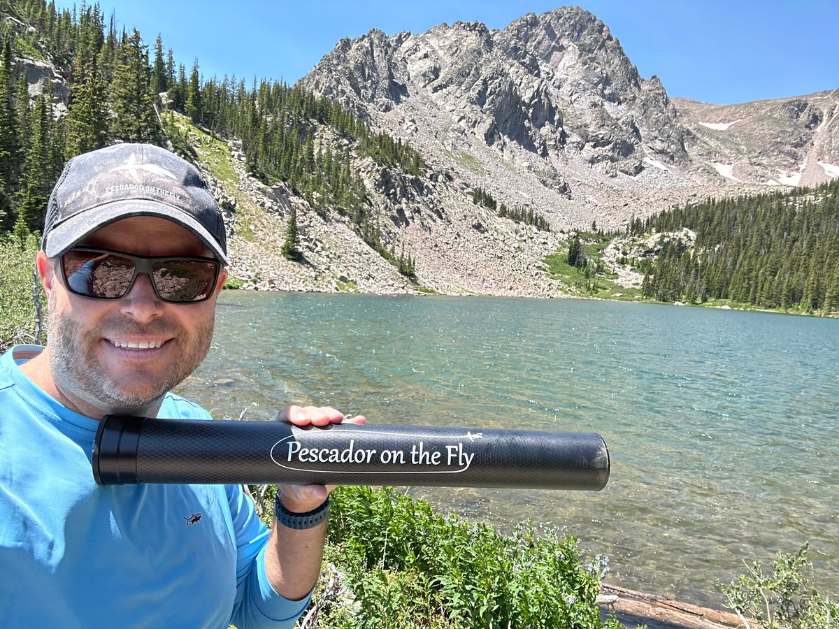 Angler holding the short black Pescador on the Fly carbon rod tube at the shore of an alpine lake below rocky peaks